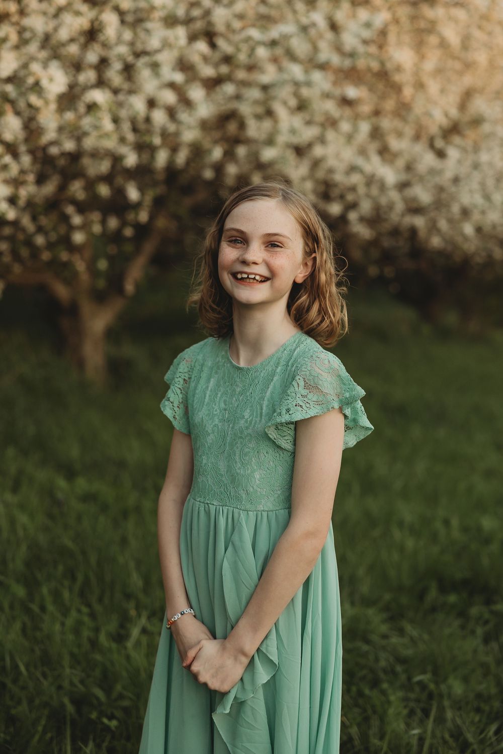 Smiling girl in mint green lace dress standing outdoors near blooming trees on green grass.