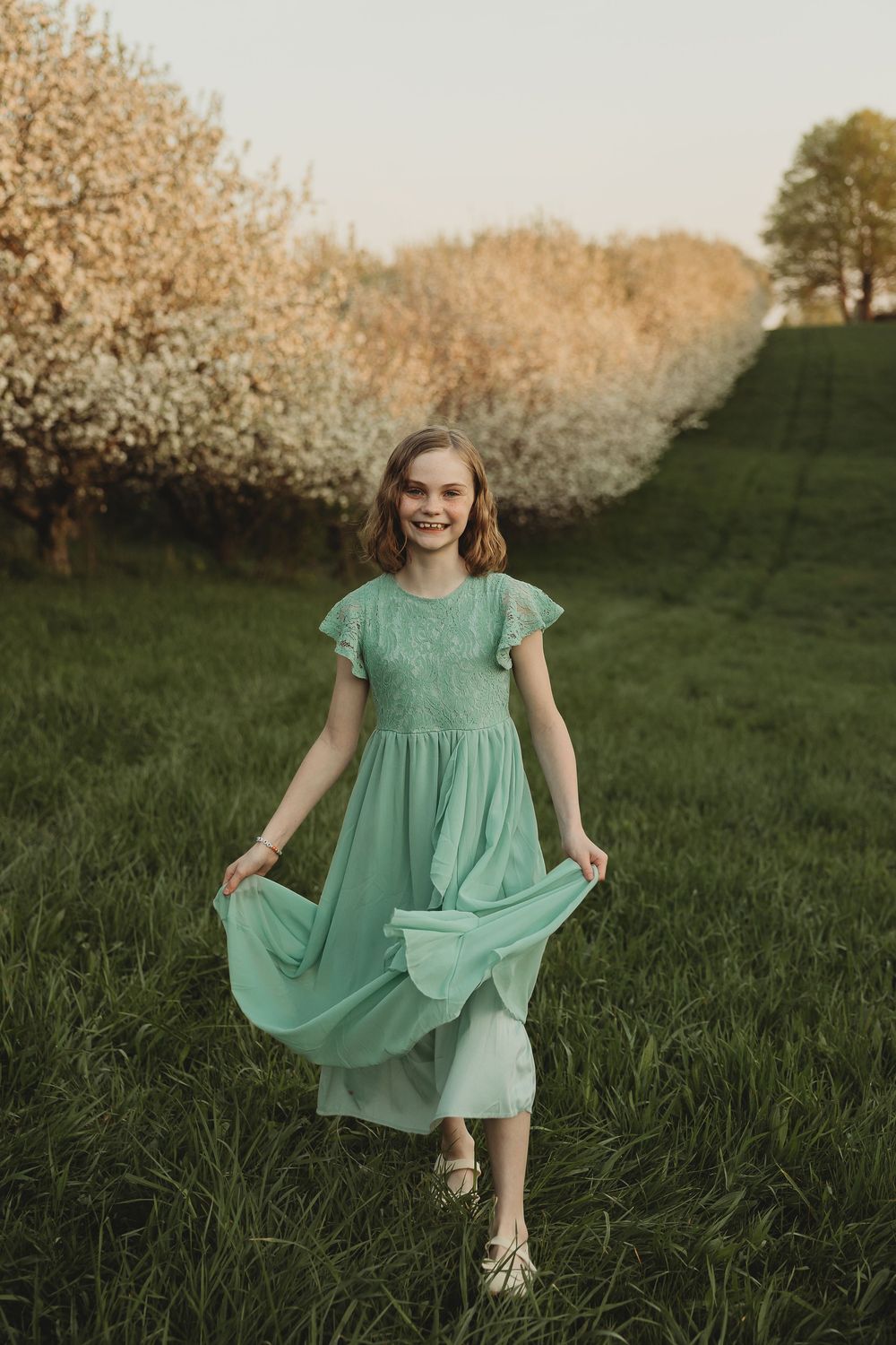 Young girl in mint chiffon dress holding skirt while standing in green field with flowering trees.