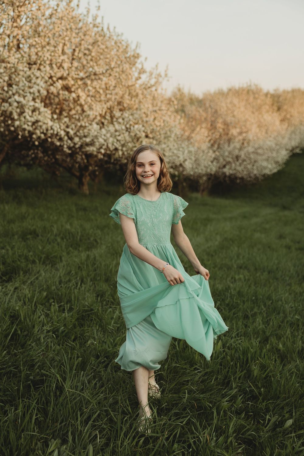 A smiling girl in a mint green dress walks barefoot through lush grass near blooming trees at dusk.