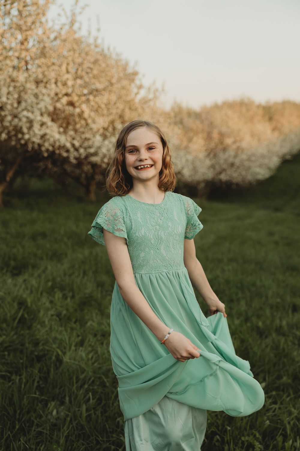 Smiling girl in mint green lace dress twirling in blooming orchard during golden hour.
