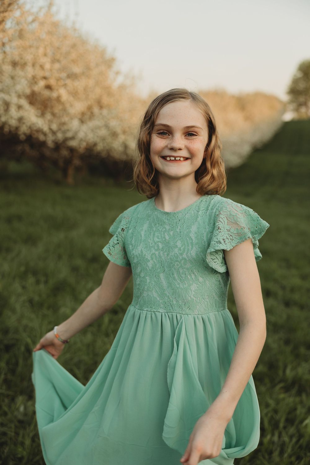 Young girl in mint green lace dress smiling outdoors among blooming trees on green grass.