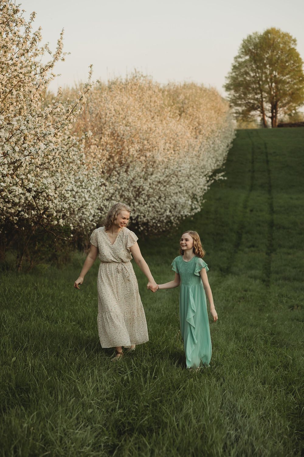 Mother and daughter in elegant dresses holding hands in a blooming orchard on a sunny spring day.