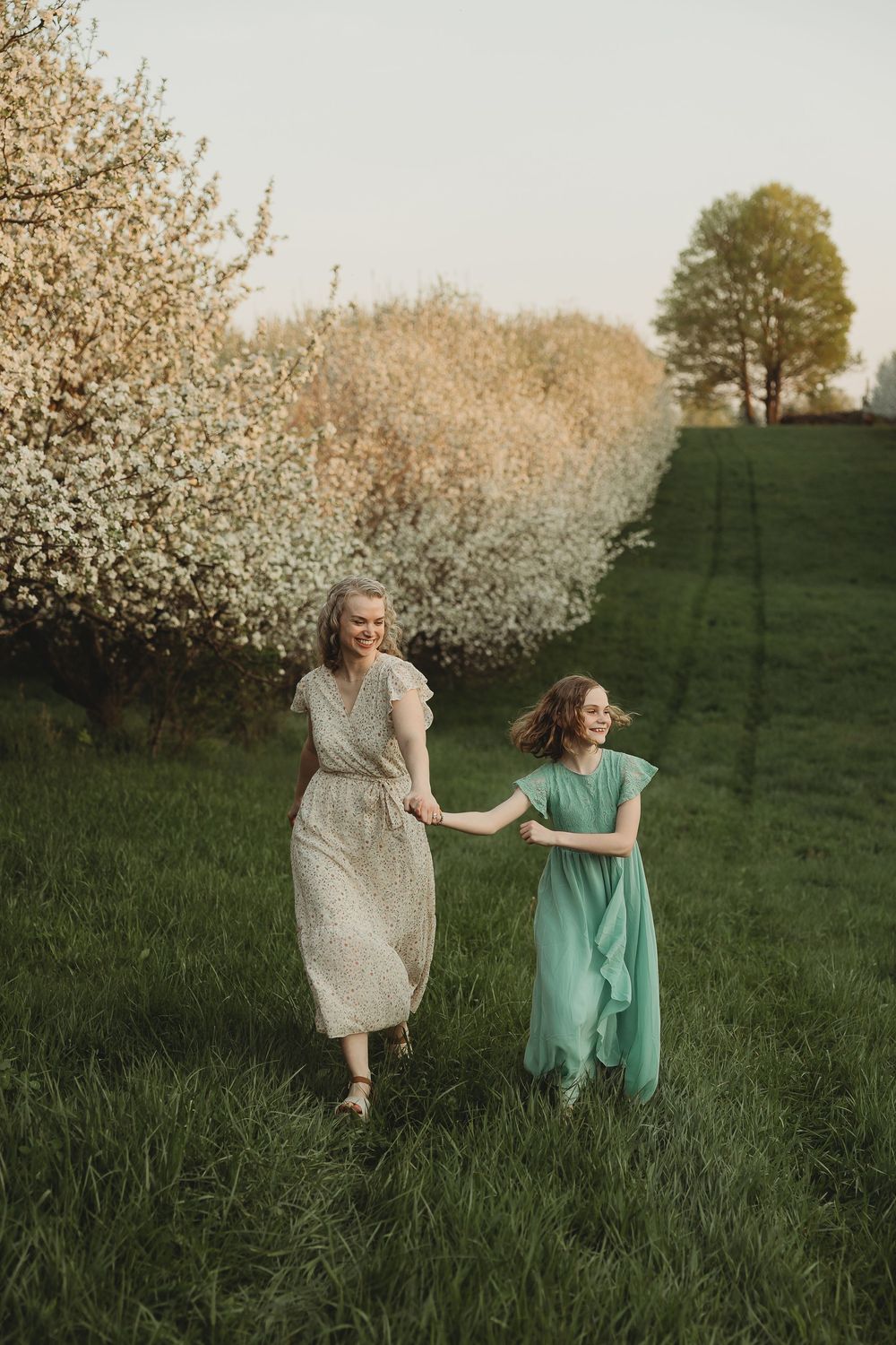 Two women in elegant dresses hold hands while walking through a blooming orchard on a sunny spring day.
