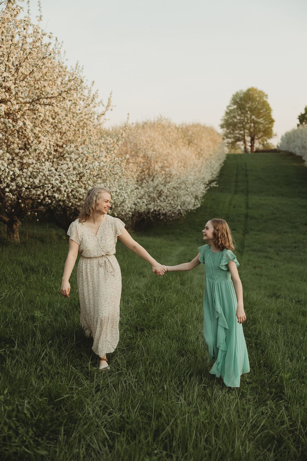 A mother and daughter in elegant dresses hold hands while walking through a blooming orchard on a lush green hillside.