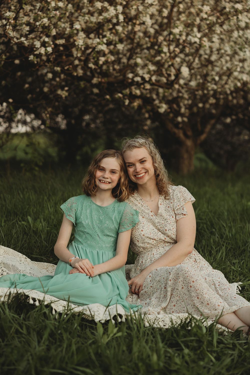 Two smiling girls in floral and teal dresses sit on a blanket in green grass beneath a blooming white flowering tree.
