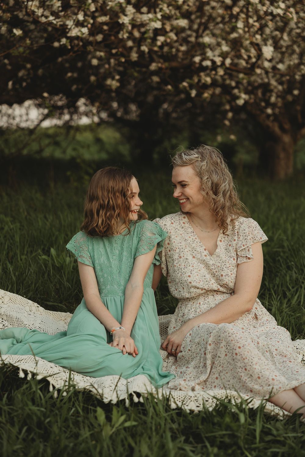Two women in floral dresses sitting on a blanket in green grass, smiling at each other near blooming trees.