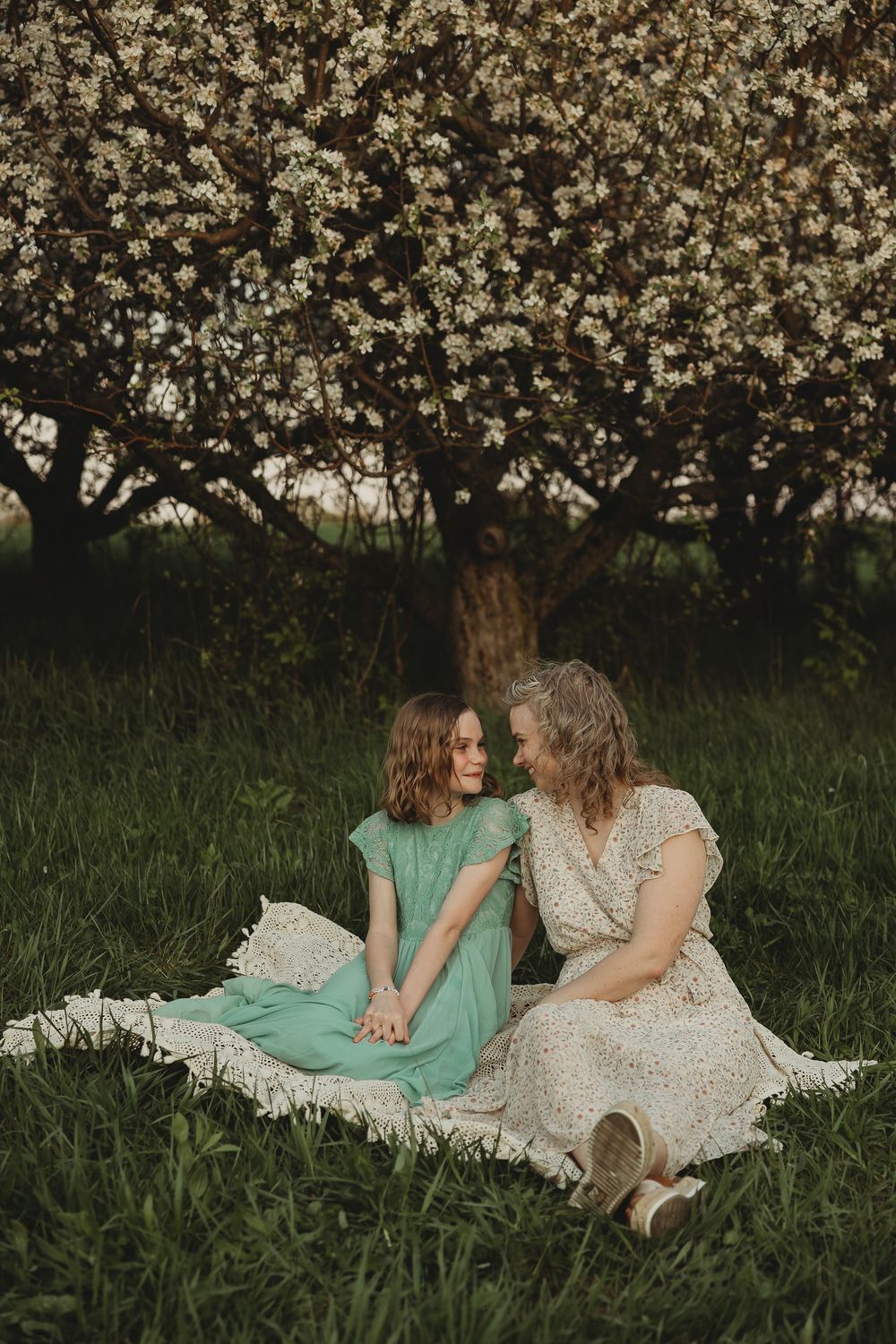 A mother and daughter sit together on a blanket beneath blooming spring trees in a lush green garden.