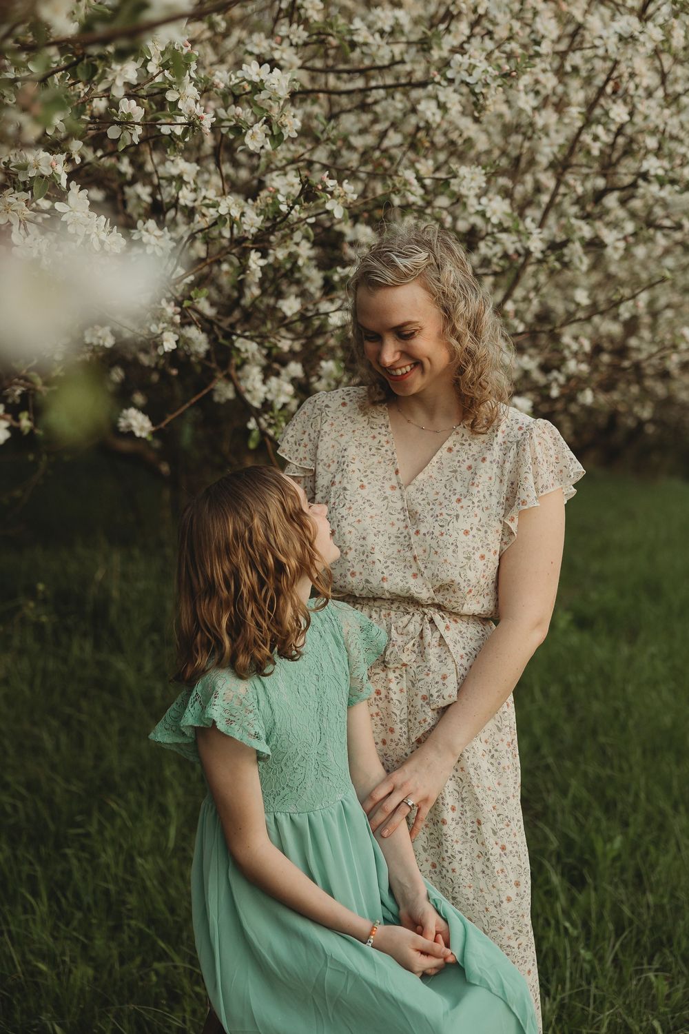 A smiling woman in a floral dress holds hands with a girl in a teal dress near a blooming white flower tree.