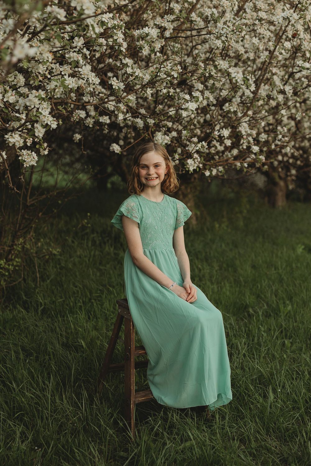 Happy girl in long mint green dress sitting on wooden stool beside white flowering tree.