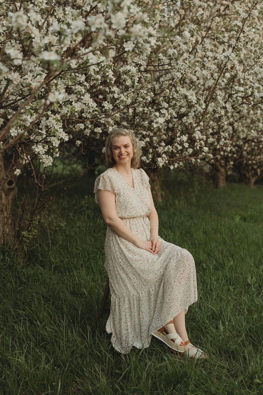 A smiling woman in a floral midi dress sits outdoors among blooming white spring trees on lush green grass.