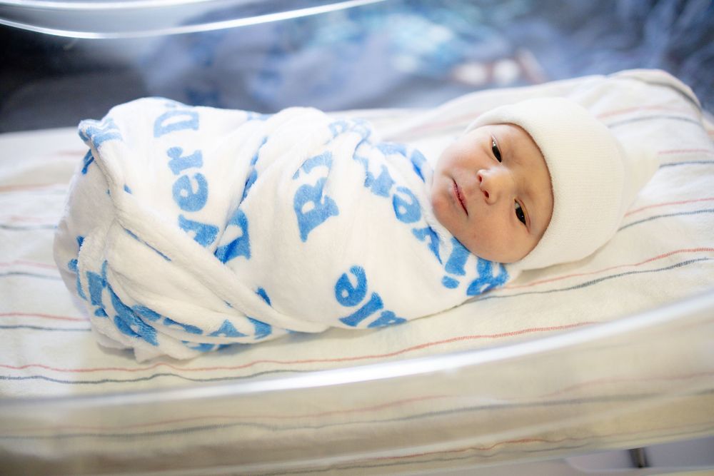 Newborn baby swaddled in blue and white blanket wearing a white hat, resting peacefully in a hospital bassinet.