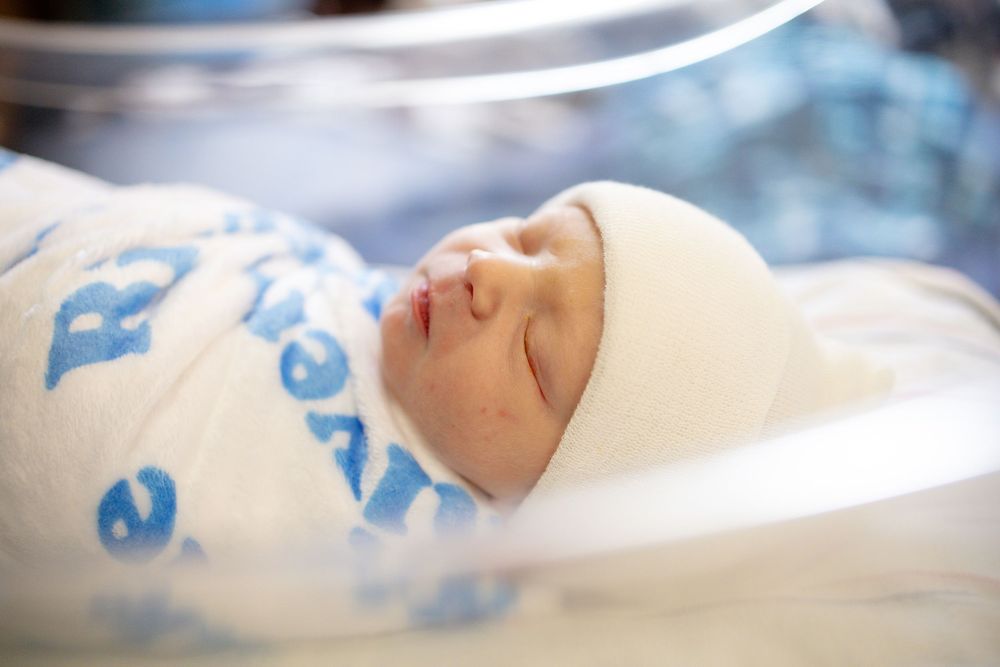 Newborn baby sleeping peacefully in hospital bassinet, swaddled in white blanket with blue letters and wearing a white hat.