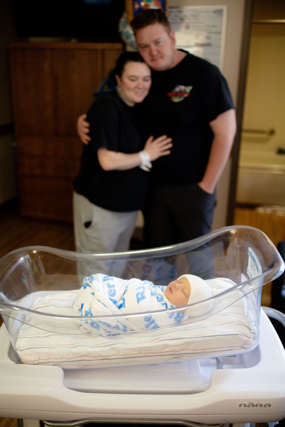 New parents embrace lovingly behind their swaddled newborn baby lying in a hospital bassinet.