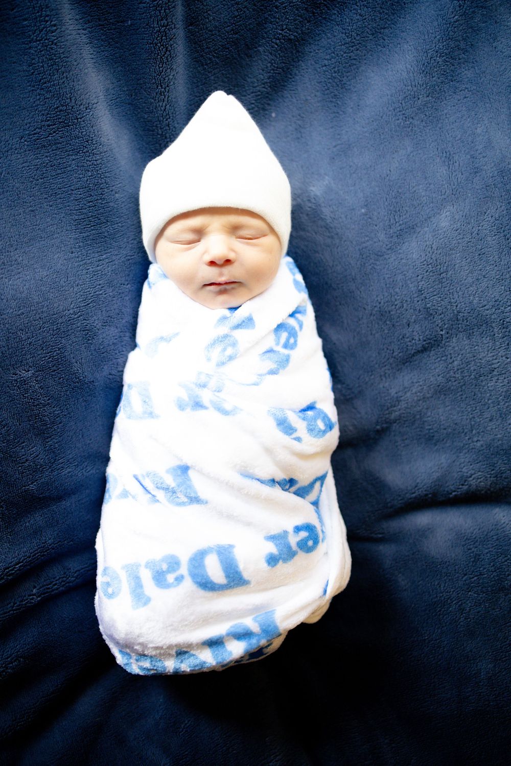 Newborn baby swaddled in a white blanket with blue text, wearing a white knit hat, sleeping peacefully.