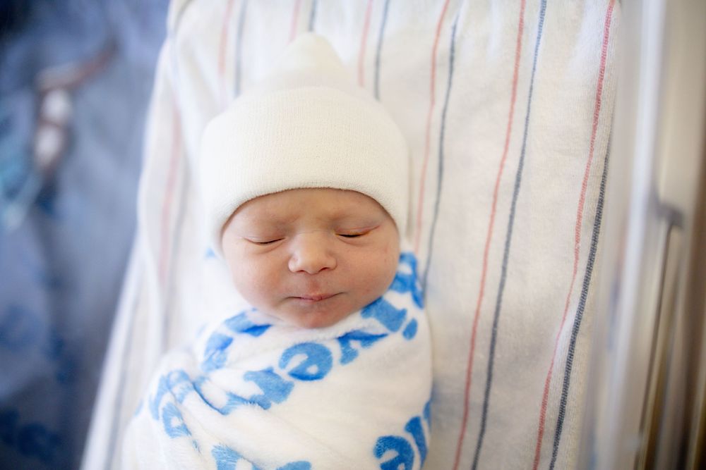 Peaceful newborn baby sleeping swaddled in white blanket with blue text, wearing a white hospital hat.