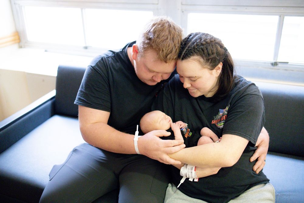 Two parents in black shirts tenderly hold their newborn baby together while seated on a couch.