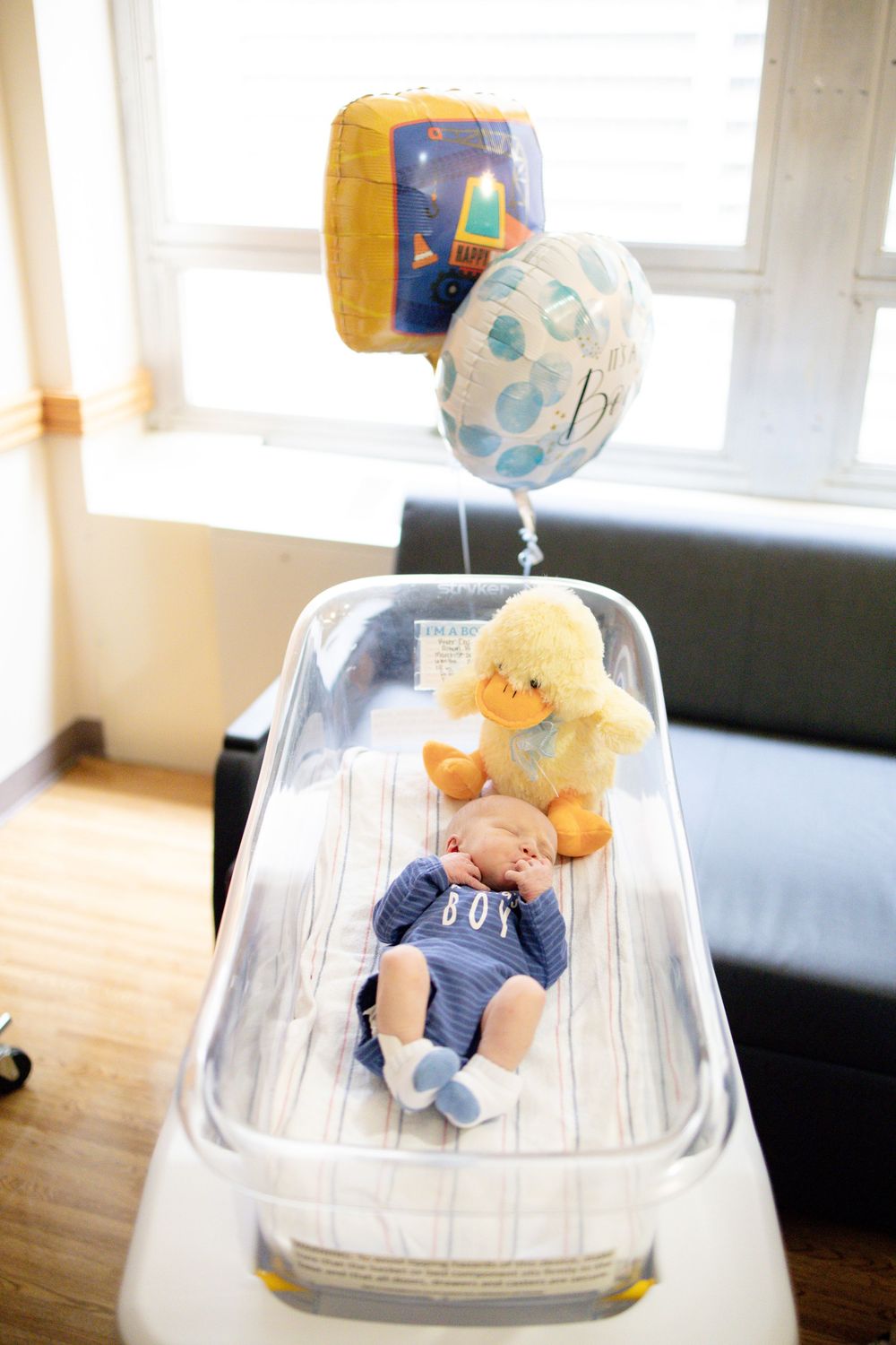 Newborn baby boy in hospital bassinet with yellow duck stuffed animal and celebratory balloons by window.