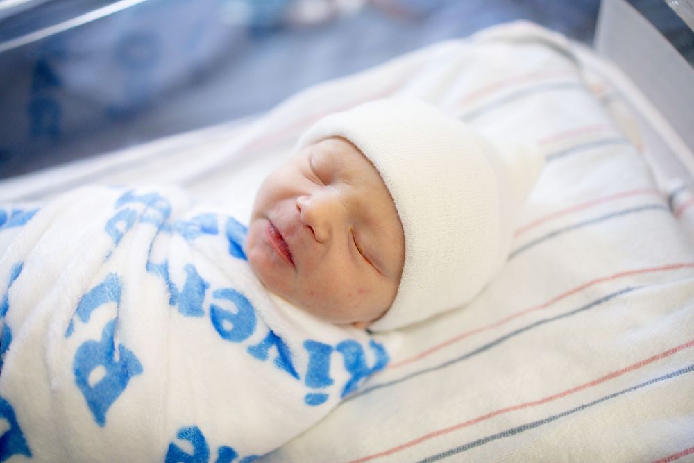 Newborn baby sleeping peacefully swaddled in a white blanket and hat inside a hospital bassinet.