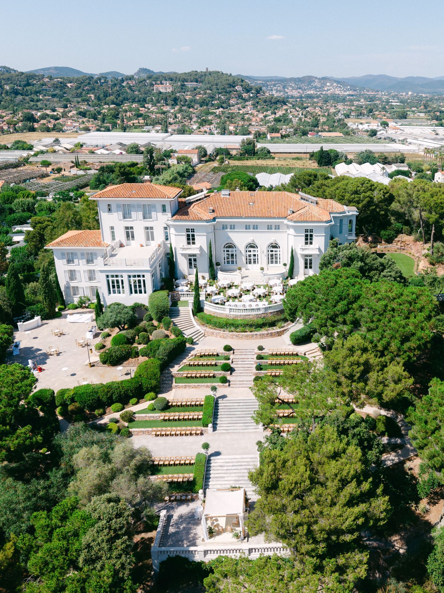 Aerial view of a grand French villa with terracotta roof, manicured gardens, and fountains surrounded by lush greenery.