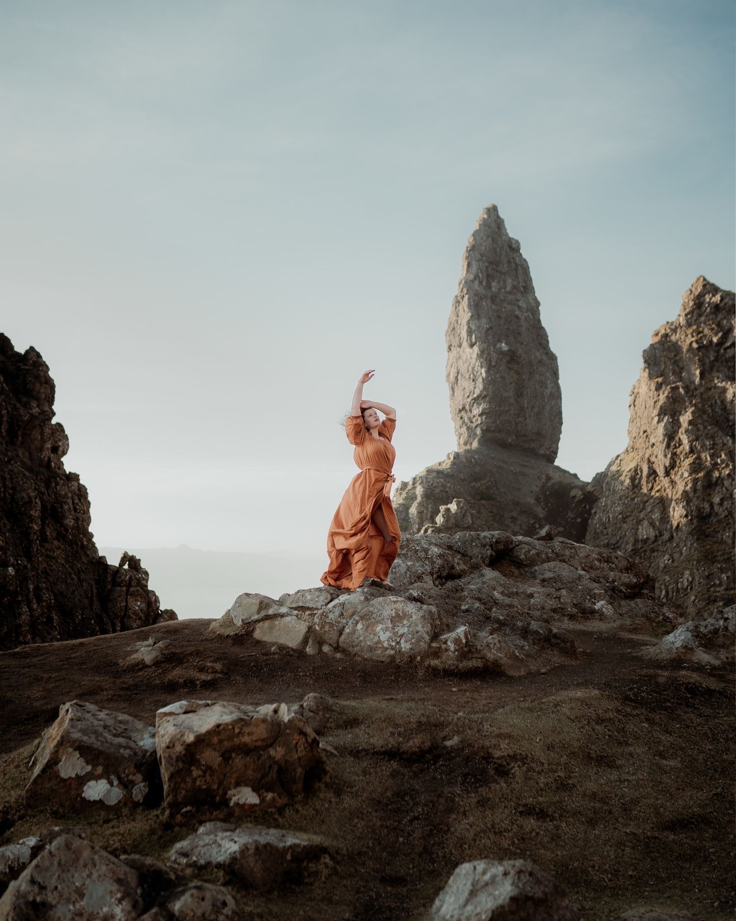 Woman in orange dress poses dramatically on rocky terrain with a towering stone pinnacle against a pale sky.