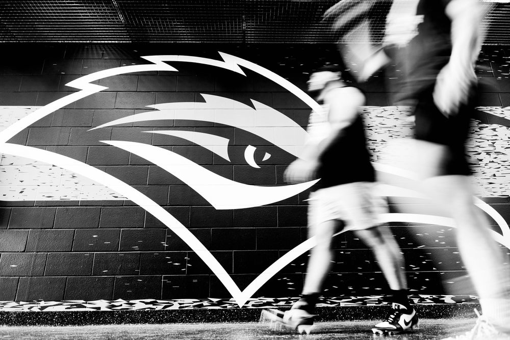 Black and white photo of athletes running past a large hawk mascot logo painted on a stadium wall.
