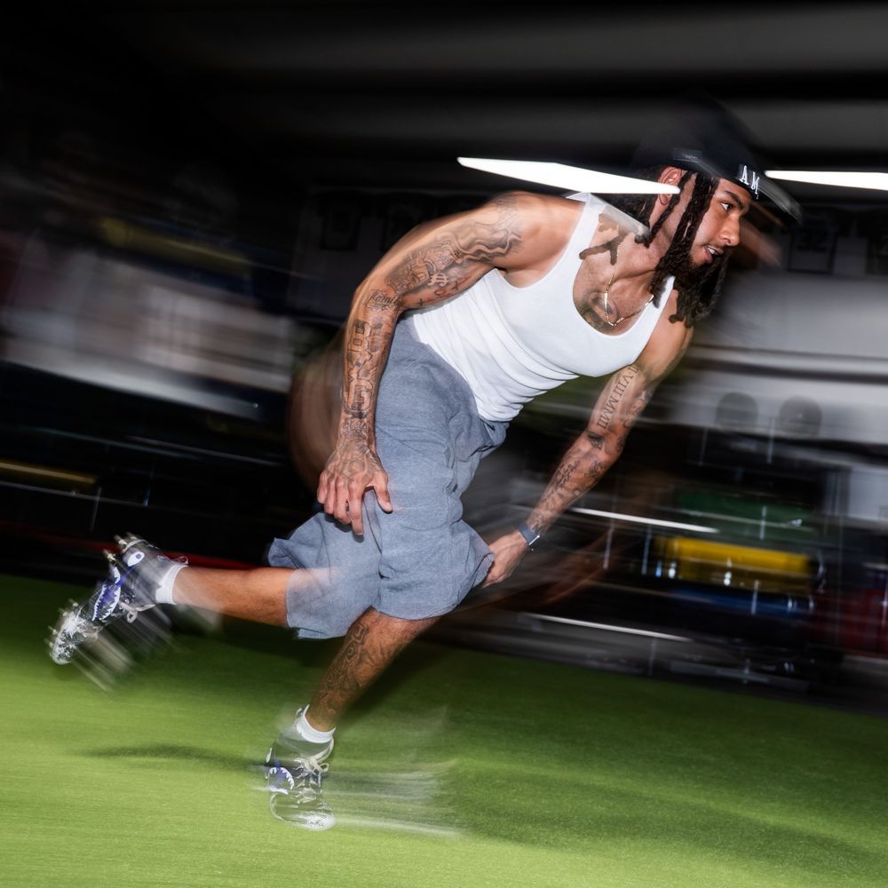 A heavily tattooed athlete sprints explosively on a turf field wearing a white tank top and baseball cap.