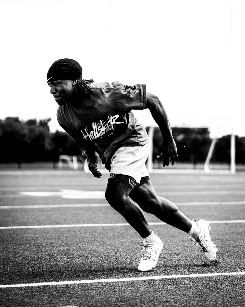 A black and white photo of a male athlete sprinting on a track wearing a Hellstar jersey and turban.