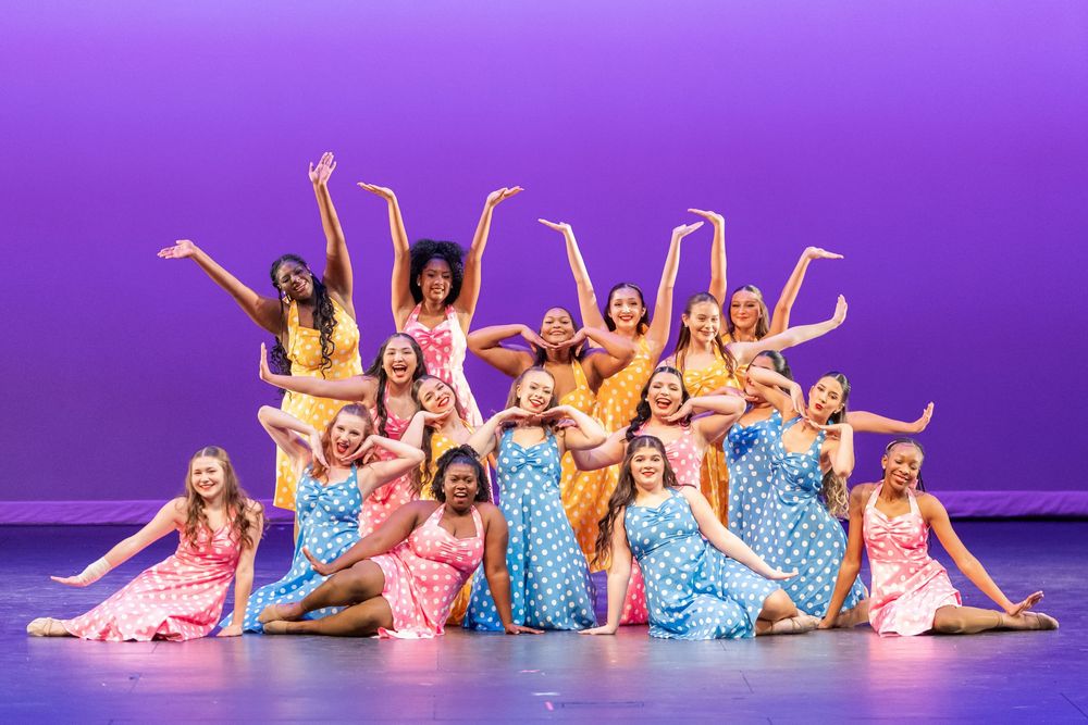 A group of smiling dancers in colorful floral dresses pose joyfully on a purple-lit performance stage.