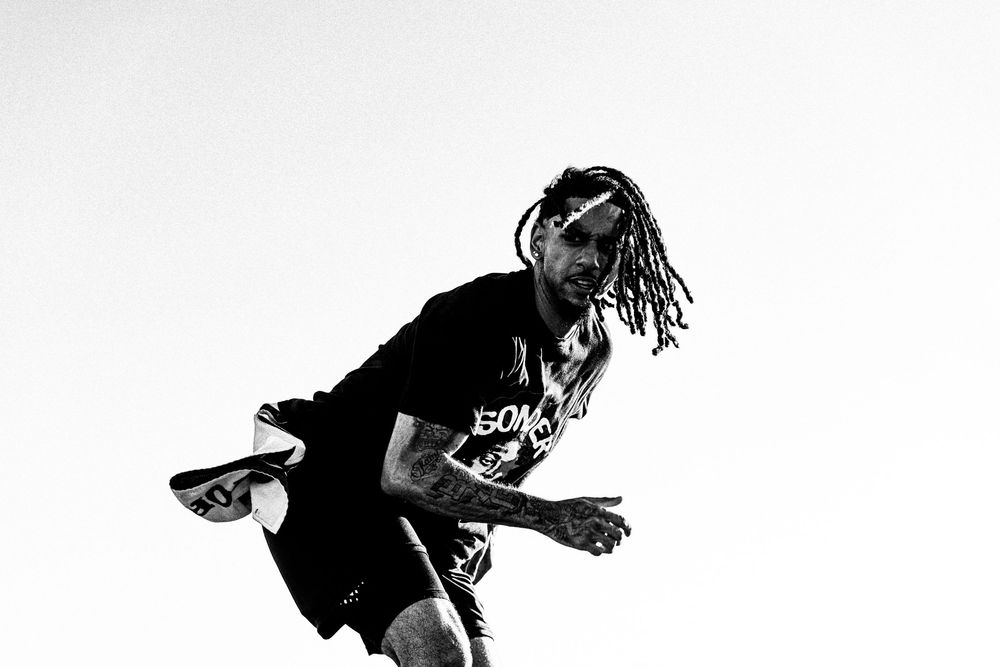 Athletic man with dreadlocks wearing black streetwear jumping dynamically in black and white photo.