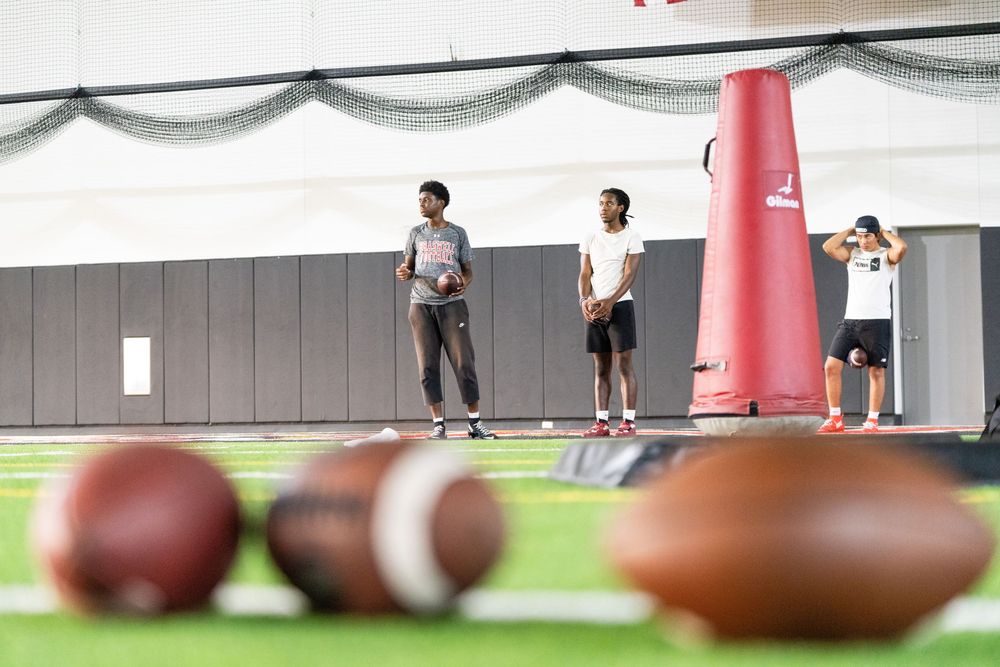 Football players training indoors on turf field with footballs in foreground and red blocking dummy.
