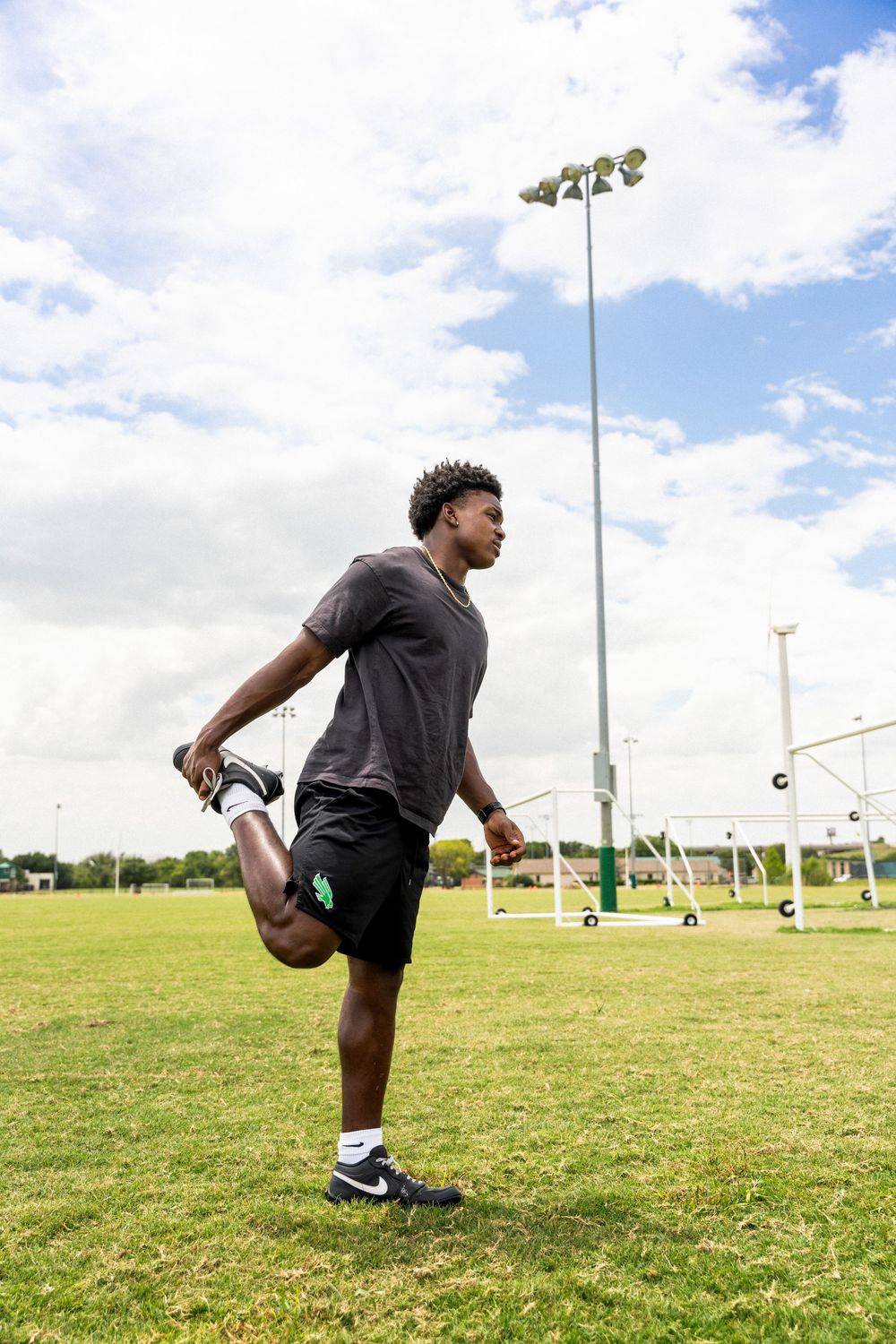 Young male athlete stretching leg on green sports field under cloudy sky wearing Nike cleats.
