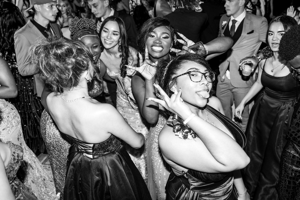 Group of teens in formal wear dancing joyfully at prom in black and white photo.