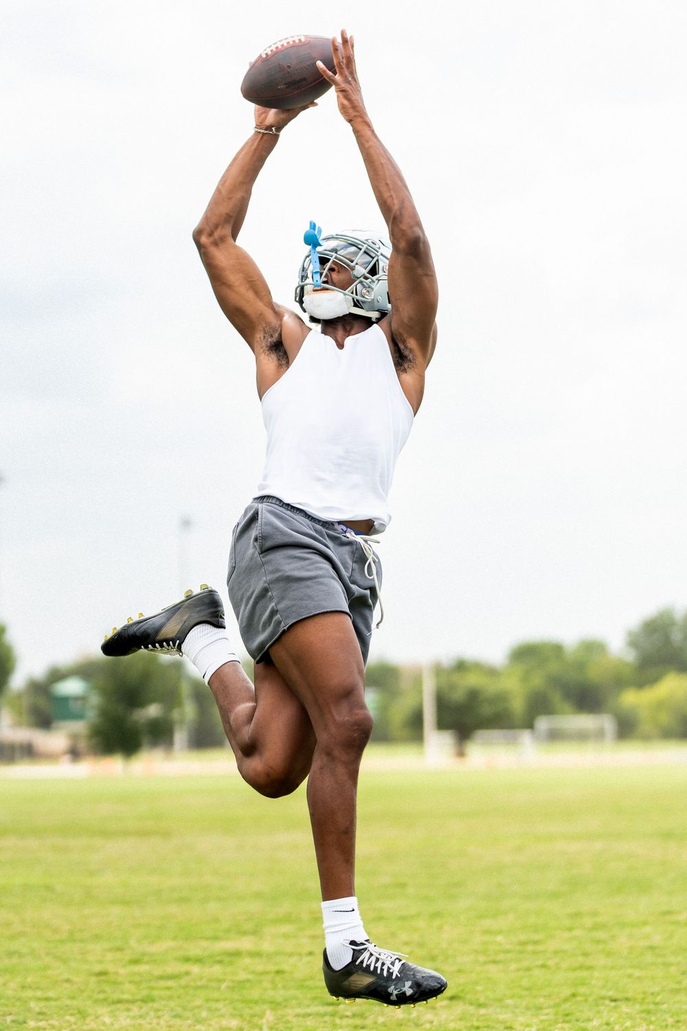 Athletic man leaping to catch football on grass field wearing helmet and white tank top.