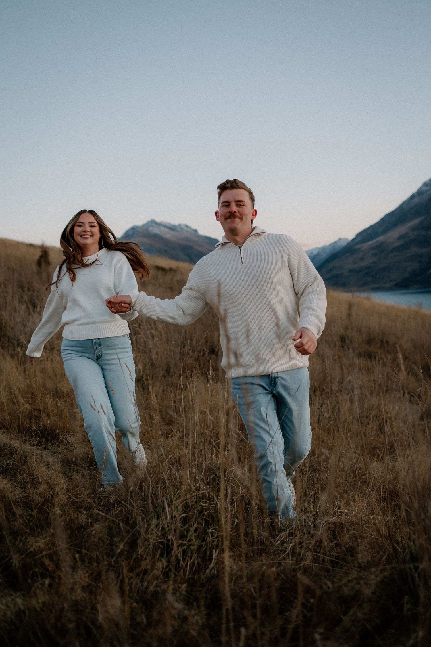 A joyful couple in white sweaters and jeans runs hand-in-hand through a golden field with mountains behind them.