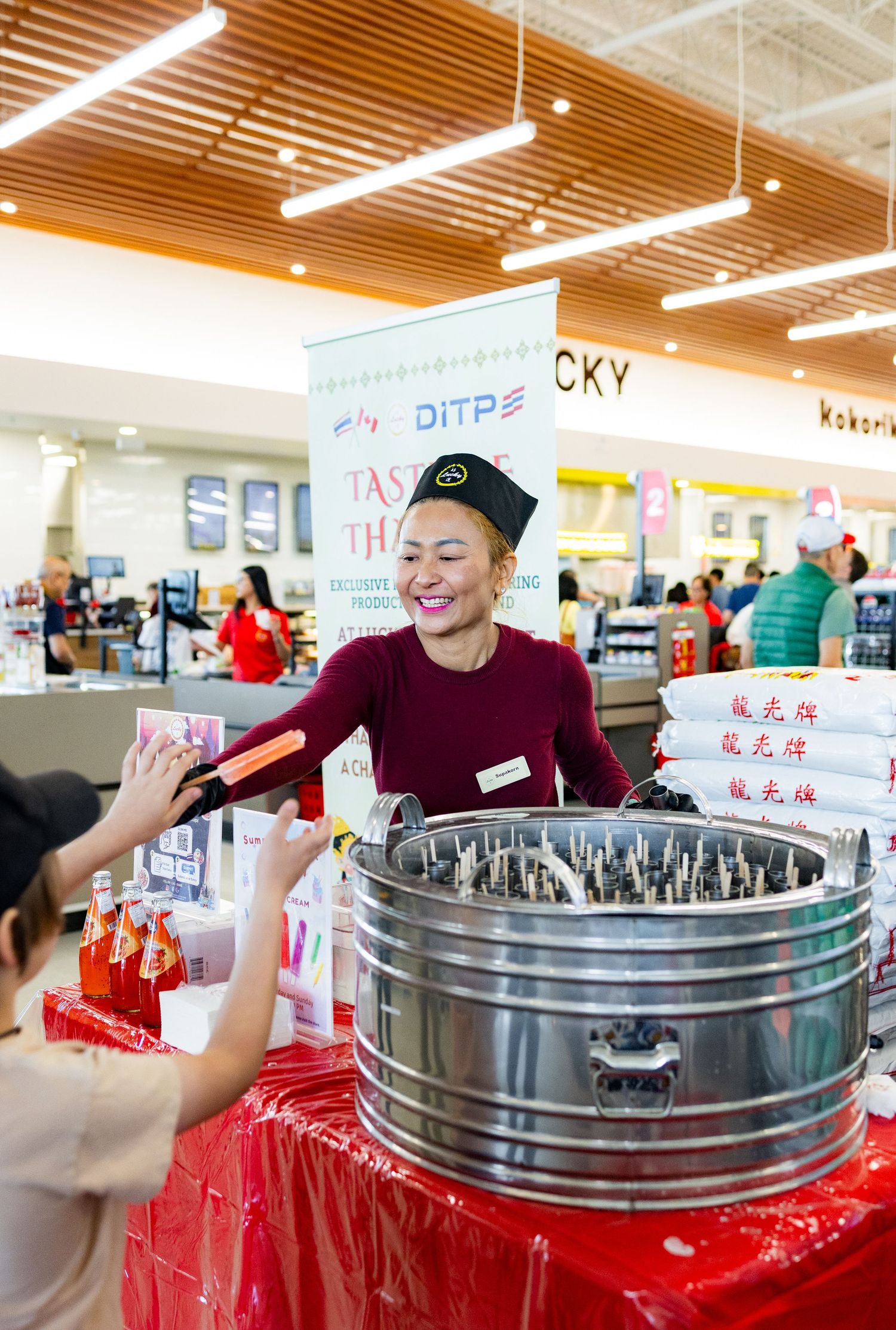 Smiling vendor in black uniform serves customers at a DITP tasting booth in a busy Asian grocery store.