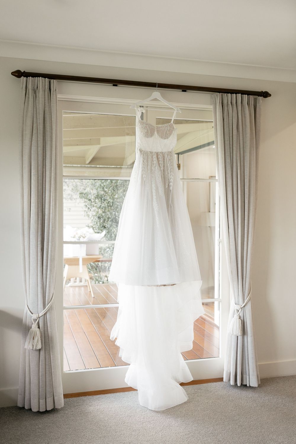 White wedding dress with lace bodice hanging on curtain rod in front of a sunlit glass door.