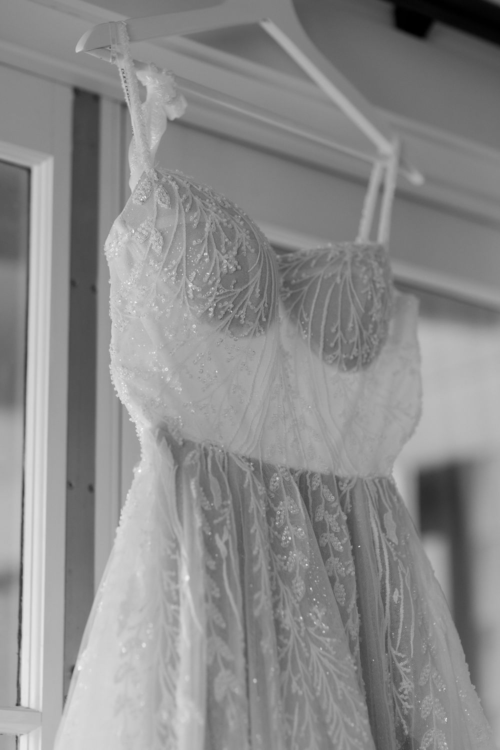 Close-up black and white photo of an elegant beaded lace wedding gown hanging on a white hanger.