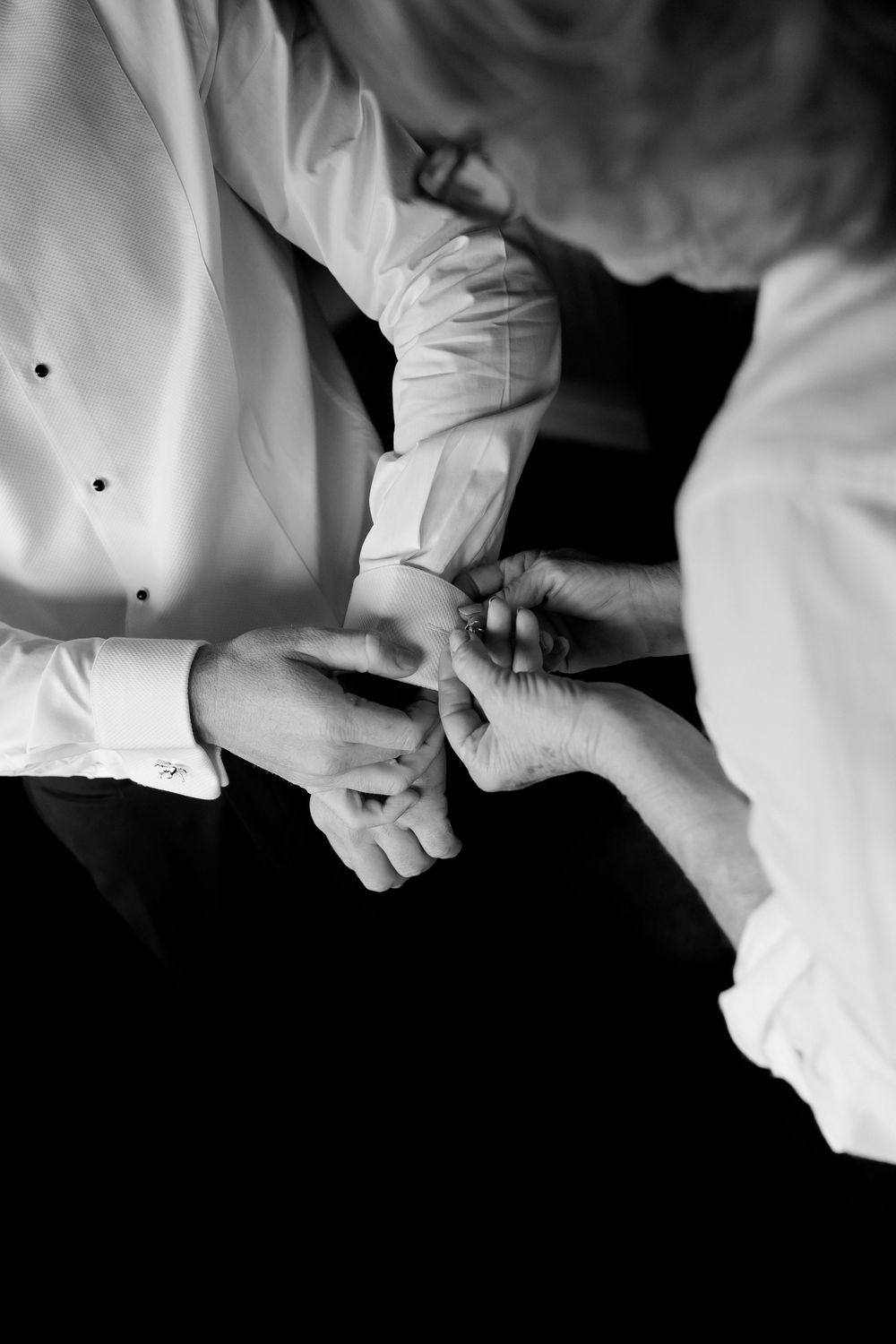 Black and white close-up of someone fastening cufflinks on a groom's white dress shirt before the wedding.