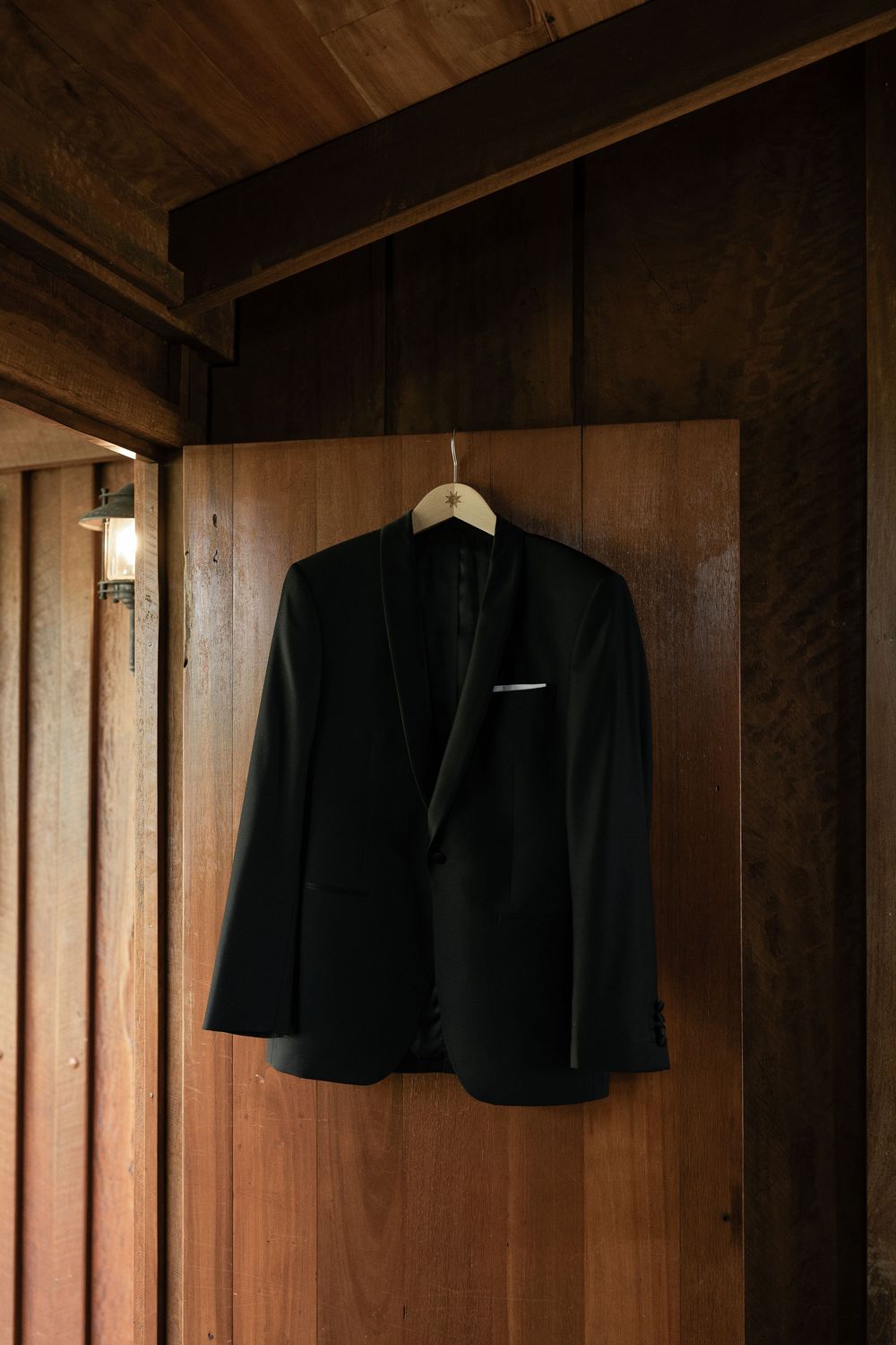 Black tuxedo jacket with white pocket square hanging on a wooden hanger against a rustic wood wall.