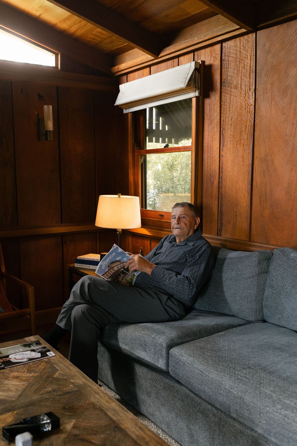 Elderly man relaxes on a grey sofa in a warmly lit wood-paneled room reading a magazine.