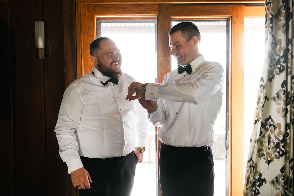 Two groomsmen in white shirts and bow ties helping each other get dressed near a window before the wedding.