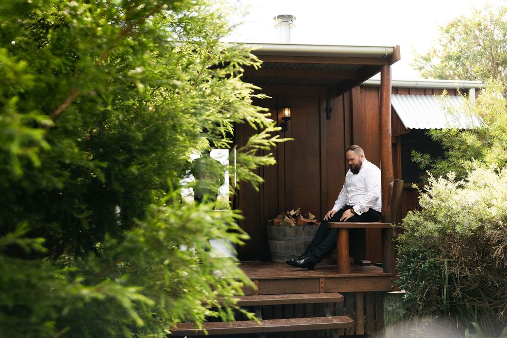 A man in a white shirt sits relaxing on a wooden deck surrounded by lush green garden foliage.