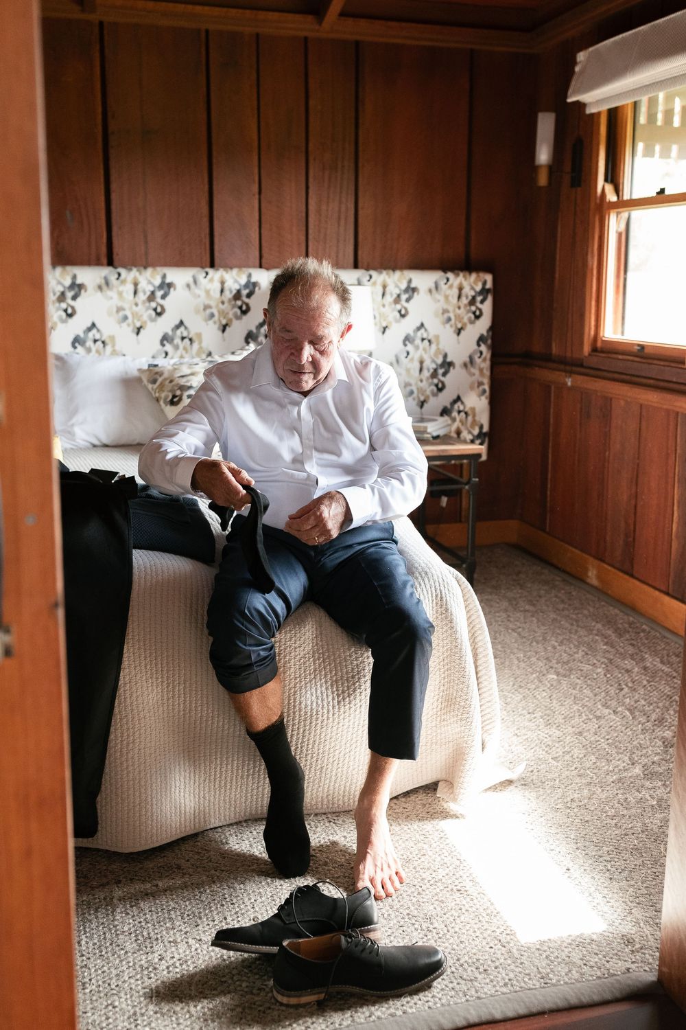 Older man in white dress shirt sits on bed in wood-paneled room preparing for a wedding ceremony.