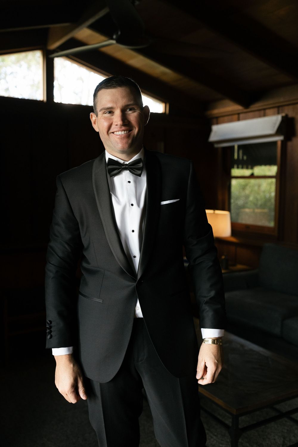 Smiling groom in black tuxedo with bow tie posing in rustic wooden venue before wedding.