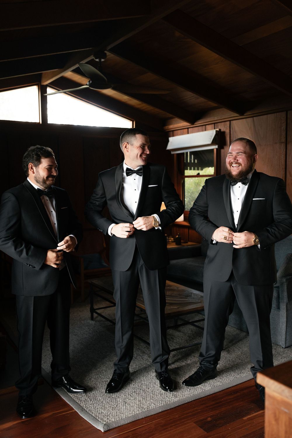 Three groomsmen in black tuxedos laughing and getting ready together in a warmly lit wooden room.