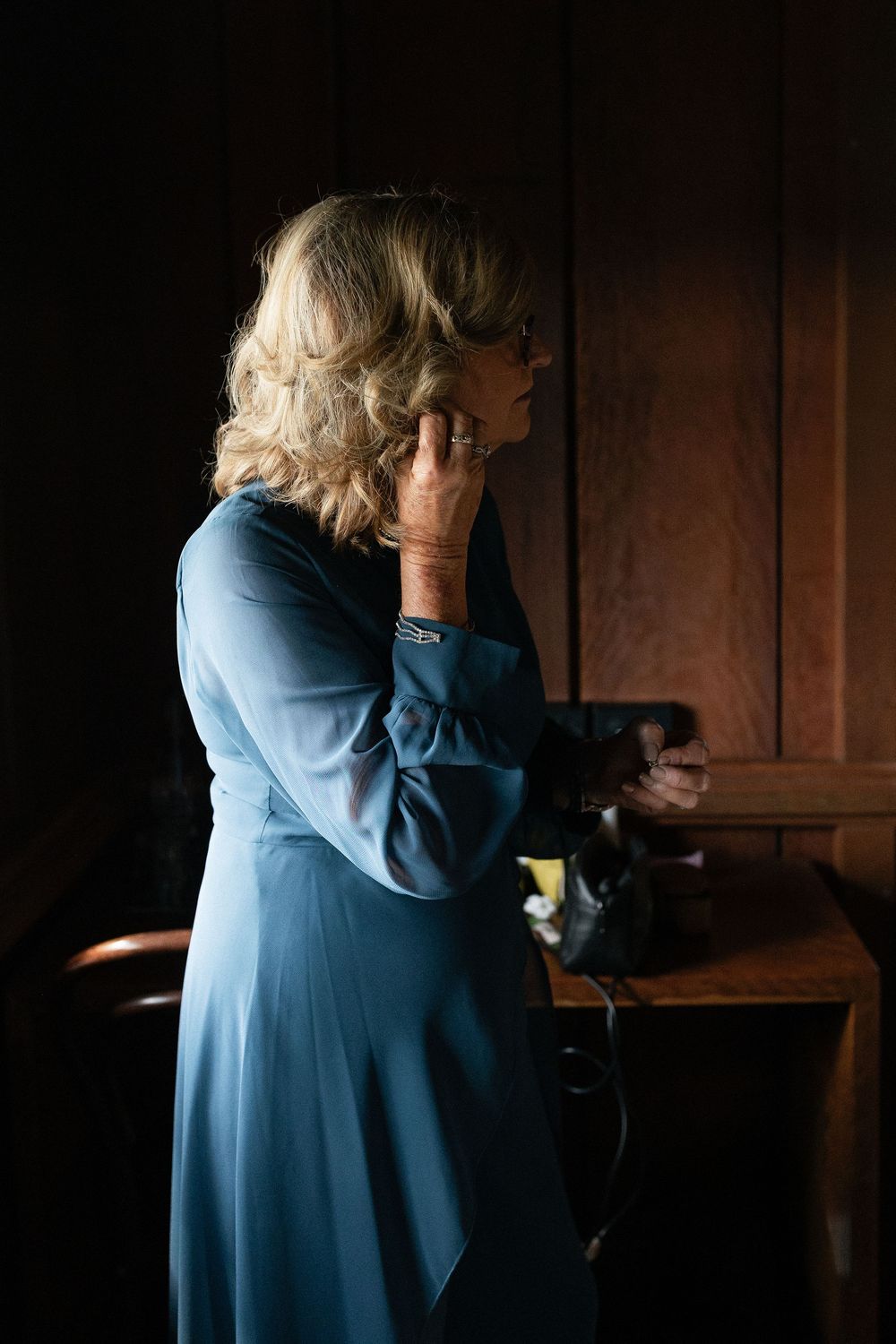 Elegant woman in blue dress putting on earrings in a warmly lit wood-paneled room before a wedding.