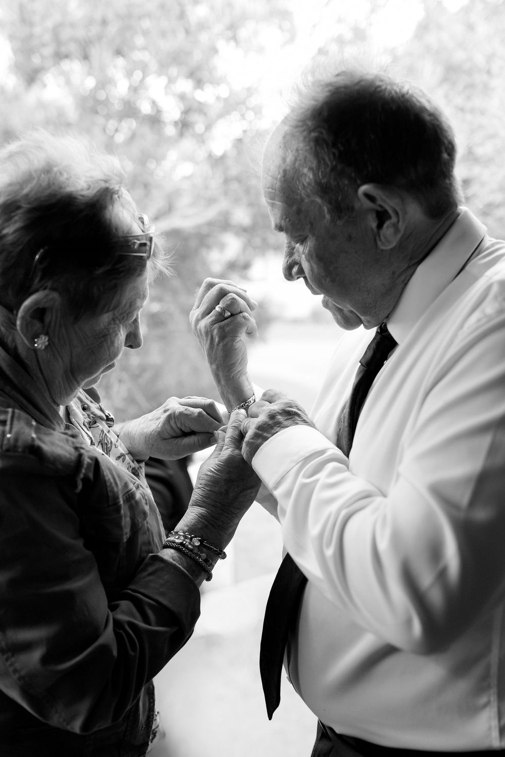 Black and white photo of elderly woman helping a man with his cufflinks near a bright window before a wedding.