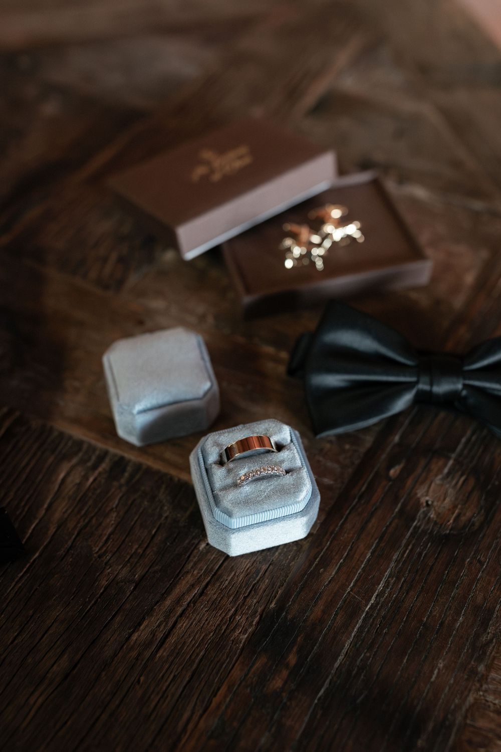 Wedding flat lay with gold rings in velvet box, cufflinks, and black bow tie on rustic wooden surface.