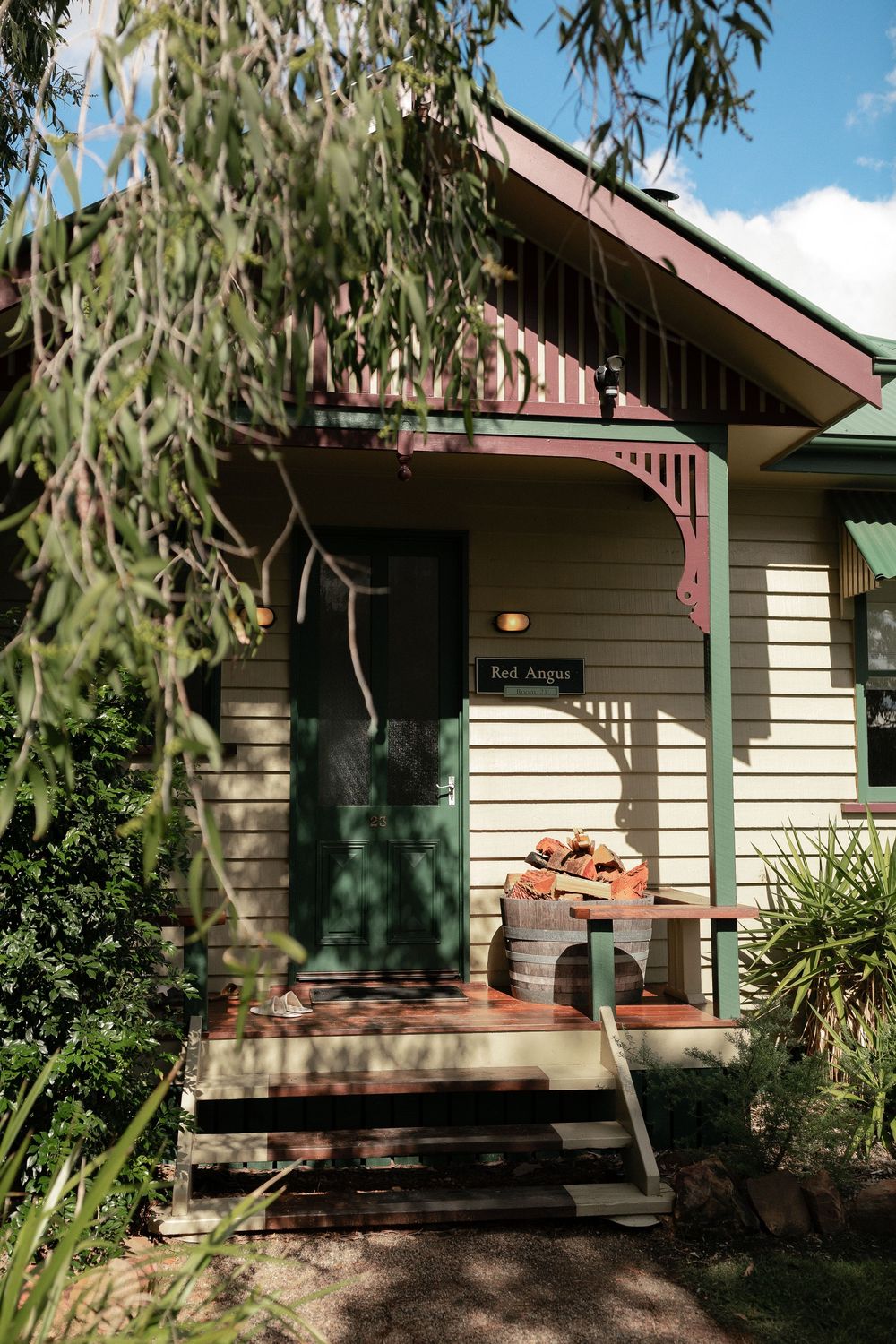 Charming Queensland cottage with green door, decorative trim, and rustic barrel planter surrounded by lush garden.