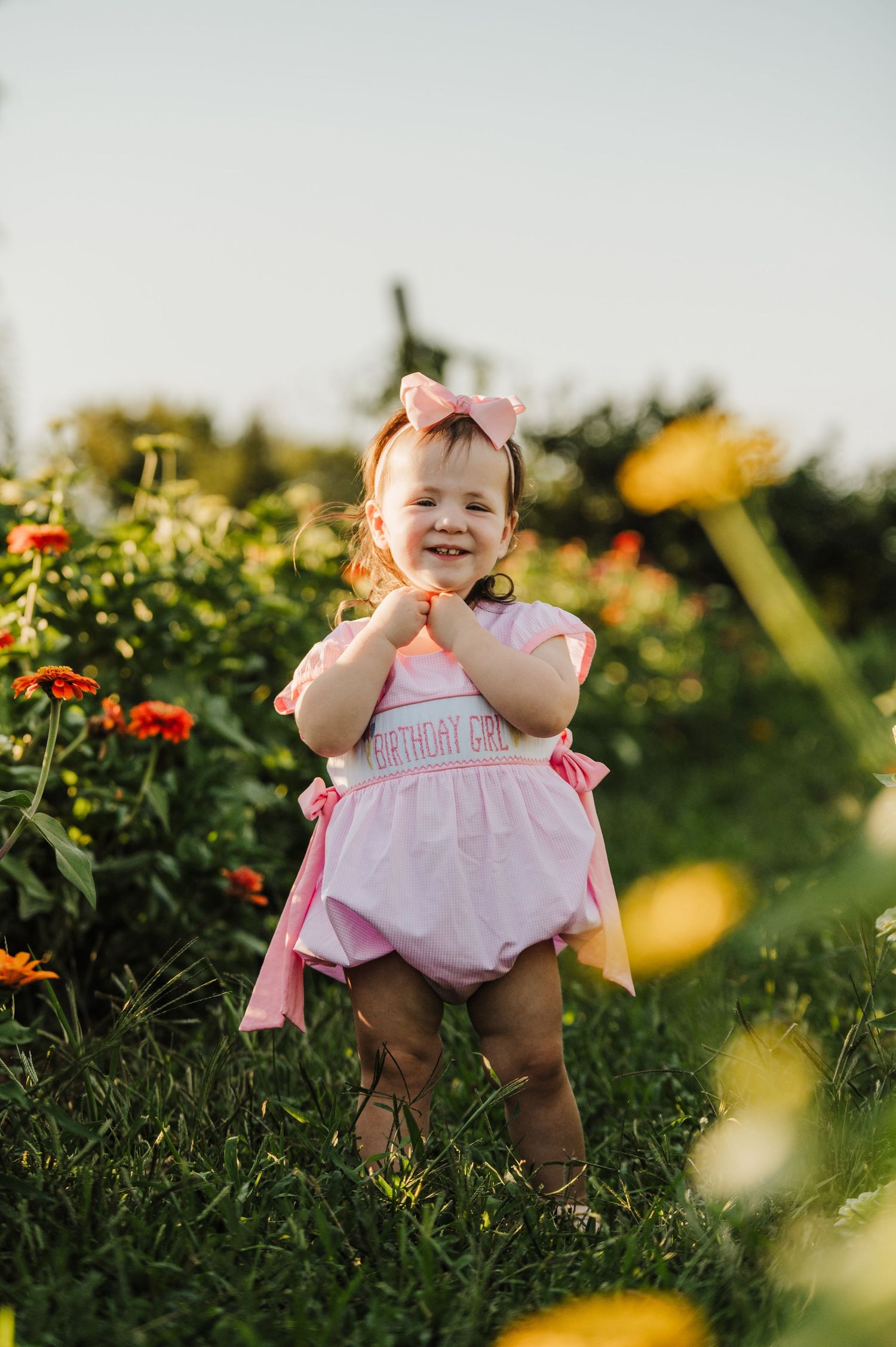Smiling toddler girl in pink birthday romper and bow headband standing in a sunny flower garden.